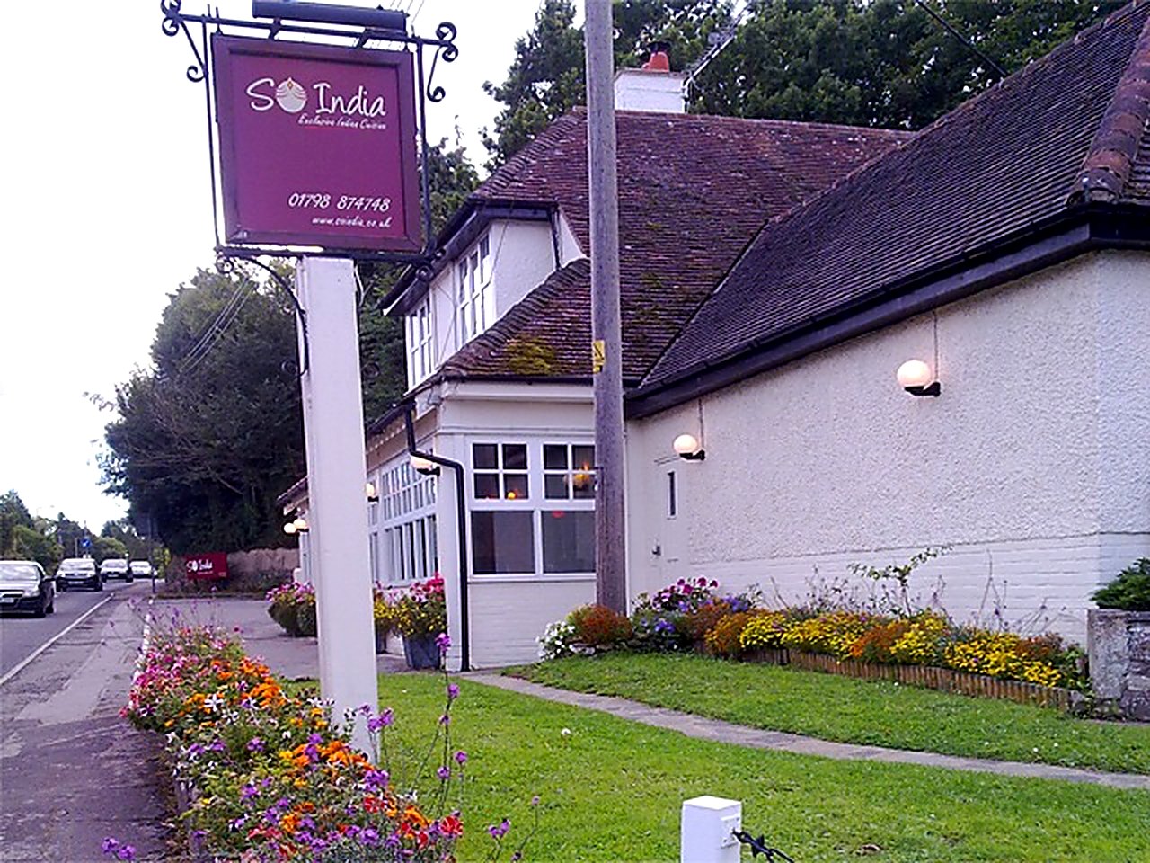 The hanging So India sign on Stane Street at dusk, with the restaurant and flower beds behind it