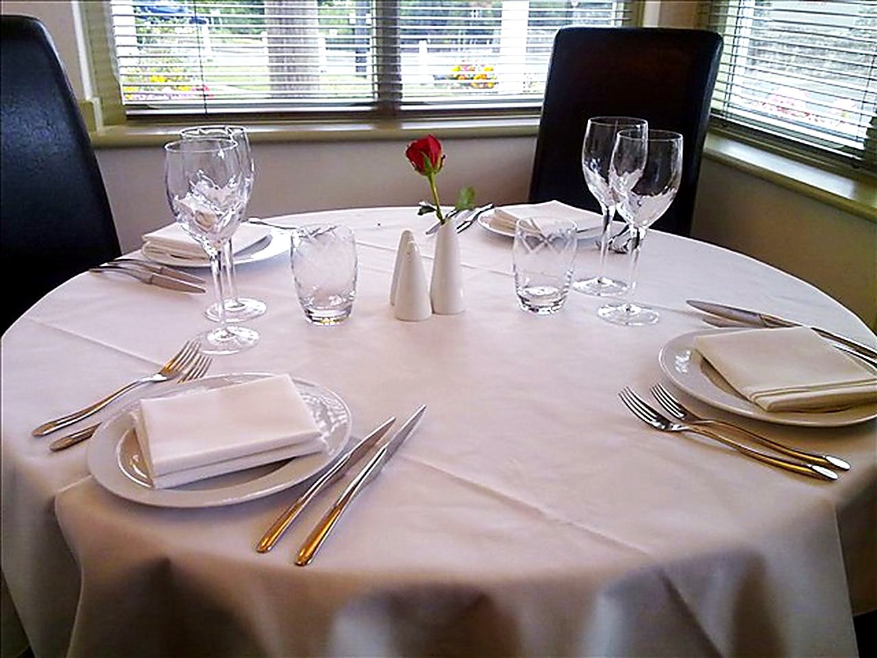 A round table set for two with wine glasses, folded napkins and a single red rose on white linen