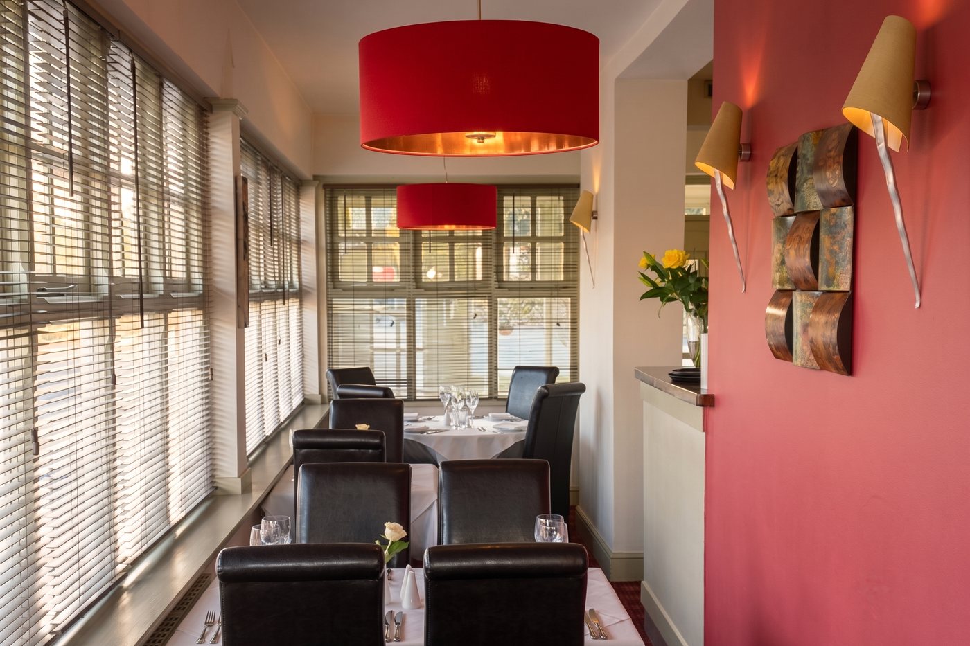 The So India dining room: white-linen tables set with wine glasses, dark leather chairs, red drum pendant lights and a deep pink feature wall
